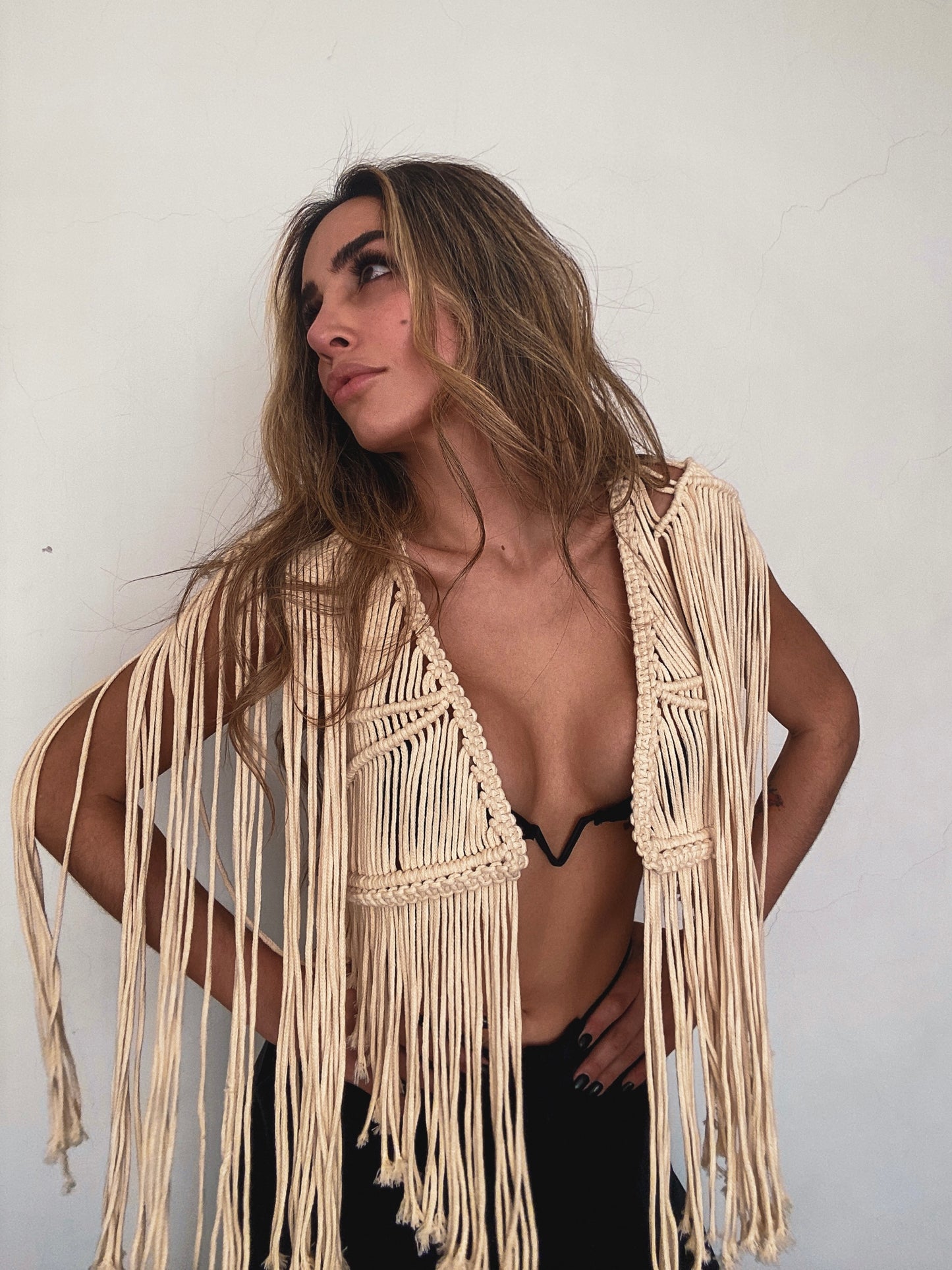 Mujer posando con actitud segura frente a pared blanca, vistiendo chaleco corto de macramé artesanal color crema con flecos largos, sobre top de bikini negro.