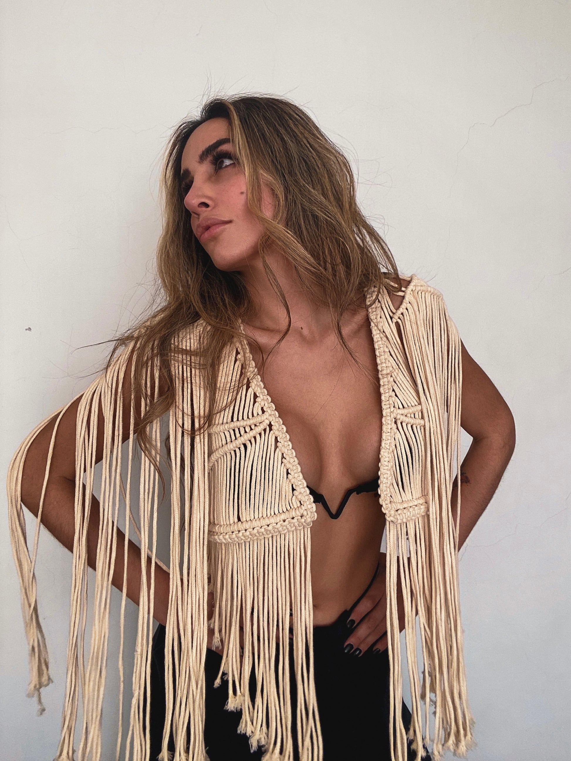 Mujer posando con actitud segura frente a pared blanca, vistiendo chaleco corto de macramé artesanal color crema con flecos largos, sobre top de bikini negro.