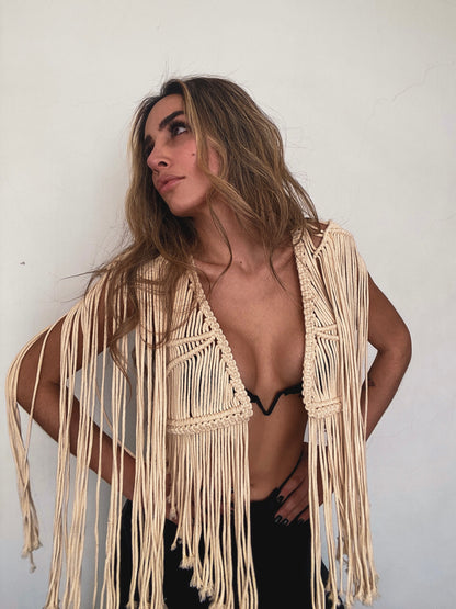 Mujer posando con actitud segura frente a pared blanca, vistiendo chaleco corto de macramé artesanal color crema con flecos largos, sobre top de bikini negro.