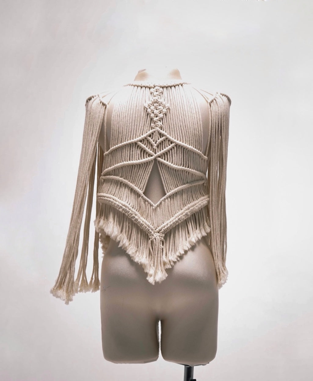 Chaqueta de macramé artesanal color crema con diseño geométrico y flecos largos, exhibida en maniquí sobre fondo blanco neutro.